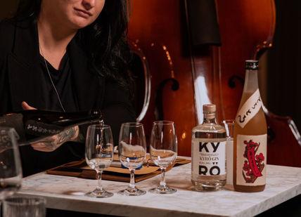 A woman with dark hair pours a drink at a table with several glasses and bottles,