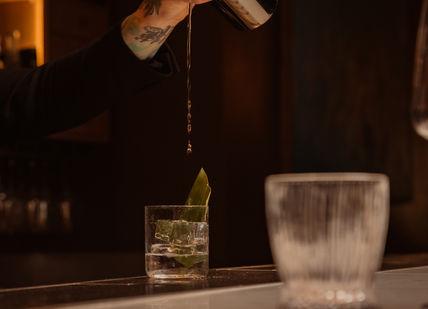 A bartender pours a drink into a glass with ice and a green leaf garnish, in a dimly lit bar.