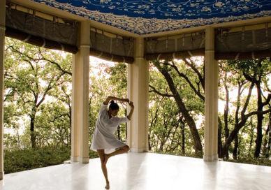 An image of a woman in a white dress standing on a porch, Yoga Retreat Programme. Ananda in the Himalayas