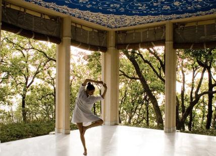 An image of a woman in a white dress standing on a porch, Yoga Retreat Programme. Ananda in the Himalayas